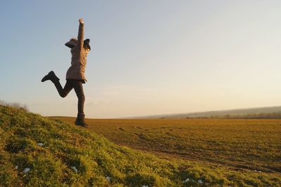 Side view of girl with arms raised running on grassy field against clear sky during sunset