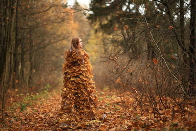 Young woman covered with leaves standing at forest during autumn