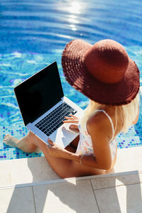 High angle view of woman wearing hat using laptop sitting by swimming pool