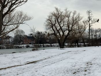 Bare trees on snow covered field by building against sky