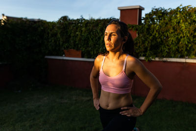 Fit female athlete in sportswear using laptop and doing lunges while following online tutorial during training in backyard at sunset