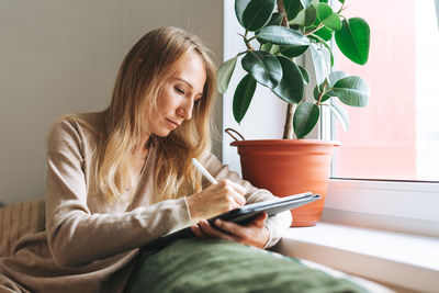 Beautiful blonde young woman artist illustrator drawing on tablet near window at home