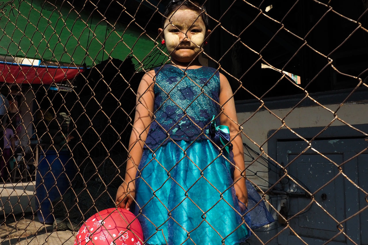 one person, real people, front view, leisure activity, lifestyles, three quarter length, fence, chainlink fence, casual clothing, blue, young adult, portrait, looking at camera, standing, metal, barrier, boundary, day, outdoors, hairstyle