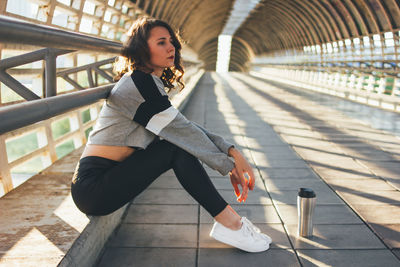 Full length of thoughtful female dancer with bottle sitting on pedestrian bridge