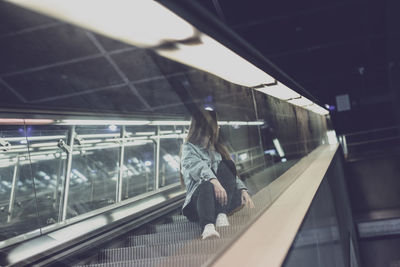 Young woman looking away while sitting on escalator seen through glass