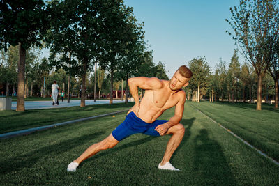 Full length of shirtless man sitting on grass against trees