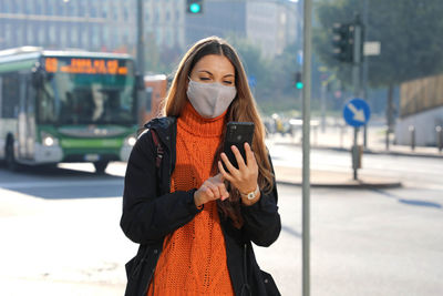 Smiling young woman wearing mask using smart phone while standing outdoors