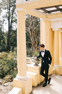 A beautiful young man, the groom in an elegant wedding suit, stands posing in the city's old park