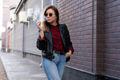 Young woman wearing sunglasses smoking cigarette while standing against brick wall