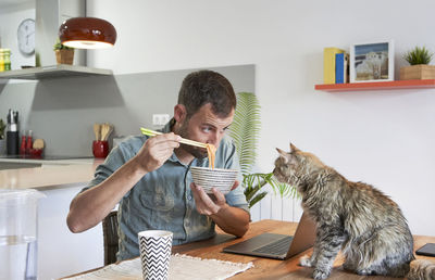 Male freelancer showing noodles to cat sitting on dining table at home