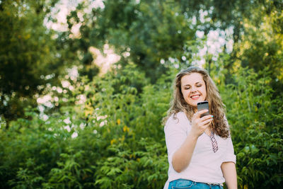 Cheerful young woman using phone against plants