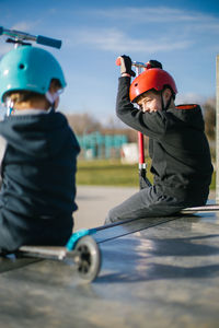 Side view of cheerful teen boy in helmet with kick scooter talking to unrecognizable friend while sitting together on ramp in skate park in sunny spring day