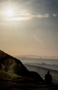 Silhouette man sitting on mountain peak against sky during sunrise