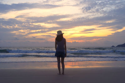 Full length rear view of woman standing at beach against sky during sunset