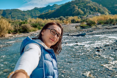 Happy, smiling woman wearing eyeglasses makes a selfie on mountains river background. 