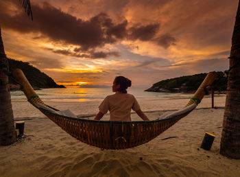 Rear view of man sitting on beach against sky during sunset