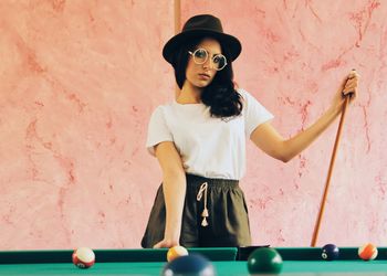 A woman wearing sexy hat and round glasses playing pool to a colorful pastel background