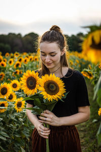 Beautiful young woman holding sunflowers while standing at farm