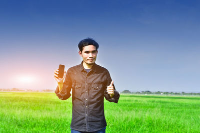 Portrait of man showing smart phone while standing on field against sky