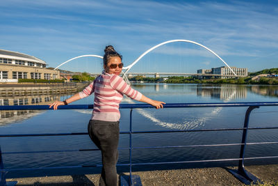 Portrait of smiling woman standing on footbridge over river against blue sky