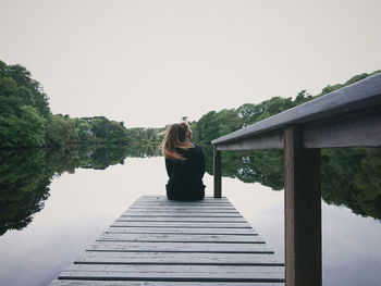 Rear view of woman sitting on pier over lake against sky