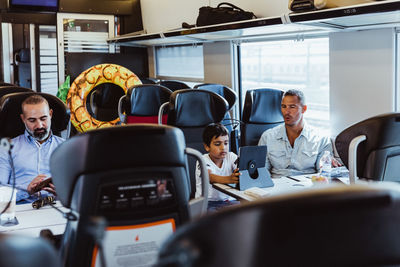 Father looking at son using digital tablet and businessman working at desk in train