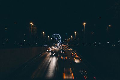 Traffic moving on road leading to ferris wheel at night