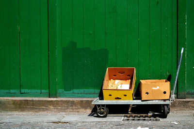 Empty boxes on push cart against wooden wall during sunny day