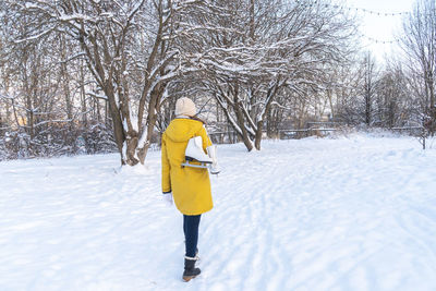 Young woman in yellow jacket with ice skates walking through snowy park