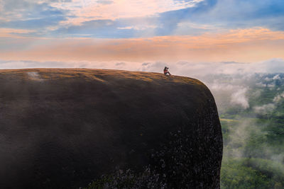 Mid distance view of woman sitting on cliff against sky during sunset