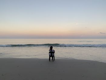 Rear view of woman sitting at beach against sky during sunset