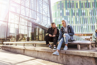 Full length of teenage friends talking while sitting on railing in city