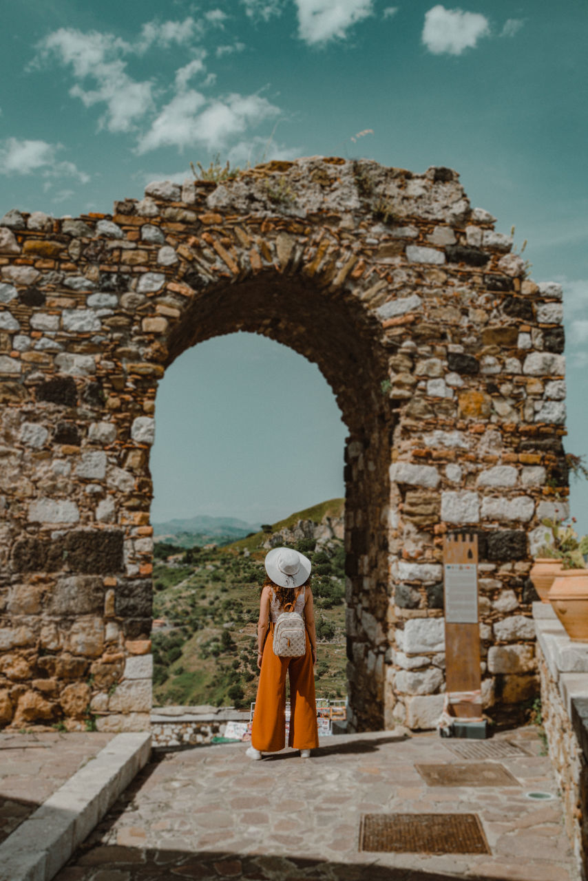 ancient history, architecture, arch, ruins, history, the past, full length, wall, sky, built structure, rock, one person, nature, old ruin, temple, ancient, adult, travel, travel destinations, clothing, standing, landmark, old, women, stone wall, day, tourism, outdoors, stone material, cloud, monument, brick, building exterior