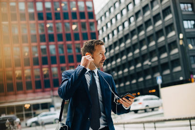 Mature businessman positioning in-ear headphones while walking against buildings in city