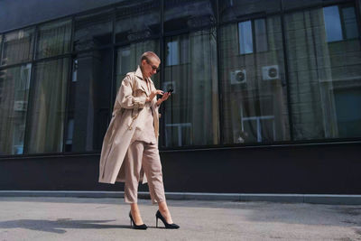 Full length of businesswoman using mobile while standing against building