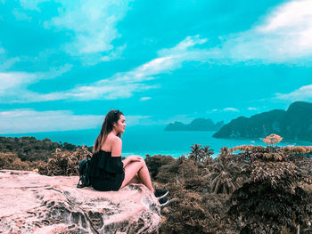 Young woman sitting on rock by sea against sky