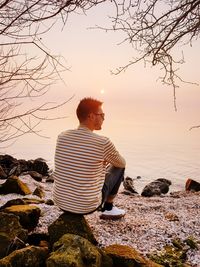 Rear view of man sitting on rock by sea against sky during sunset