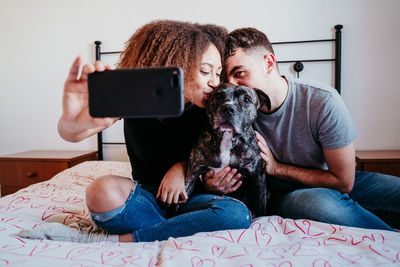 Couple taking selfie while kissing dog on bed at home