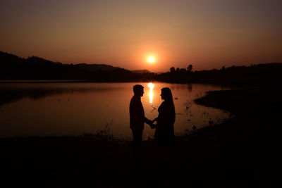 Side view of silhouette couple standing at lakeshore against sky during sunset