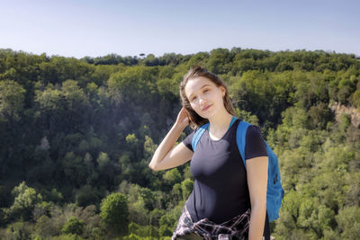 Portrait of young woman with backpack standing on mountain against clear sky during sunny day