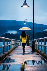 Rear view of woman with umbrella walking on pier over lake against mountains