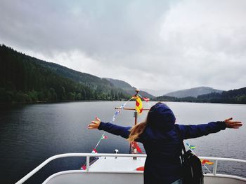 Rear view of woman with arms outstretched standing in boat on lake against cloudy sky