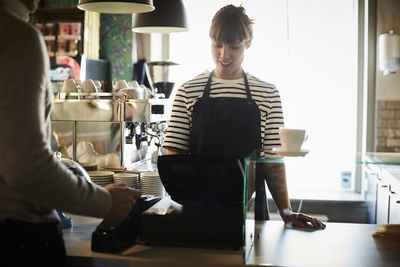 Female barista using cash register with customer in foreground at cafe