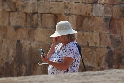 Woman wearing hat using smart phone standing against stone wall