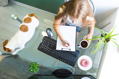 Girl sits at a glass table with a computer, holds a pen over a blank white of paper, a dog nearby. 