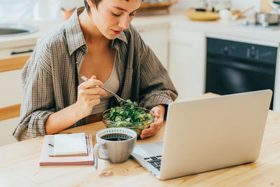 Young modern business woman eats salad while sitting in the kitchen with a computer.