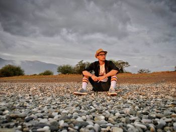 Full length of man sitting on pebbles against cloudy sky