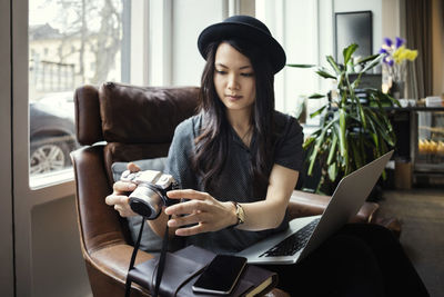 Female entrepreneur holding camera while sitting with laptop on chair