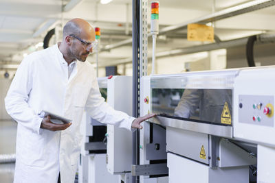 Mature male technician holding digital tablet examining machinery at factory