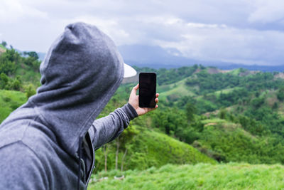 Man wearing hood taking selfie with mobile phone on mountain against sky